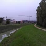 Il cantiere dei Nuovi Uffici Comunali e di Arpa. Sulla sinistra del corso d’acqua, coperto dal filare di alberi, sorge il Polo Lama Sud, complesso scolastico (nido e scuola dell’Infanzia) progettato da Giancarlo De Carlo e Associati, inaugurato nel 2008. Le linee delle due opere sembrano reinterpretare la sfrangiatura del tessuto edilizio di margine al centro urbano (© Domenico Mollura)