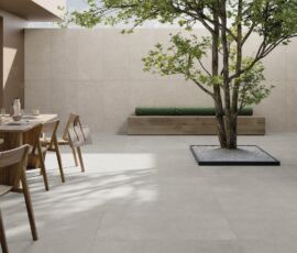 Spazio outdoor. A pavimento, finitura Grey, grigio scuro e dalla forte personalità, nel formato 60x120, spessore 20 mm da esterni. Sulla parete frontale è visibile il decoro Weave (60x120) nella variante Clay, che rievoca le texture Art Decò. La panca è rivestita con la collezione Unica, colore Beige, nel formato 20x120